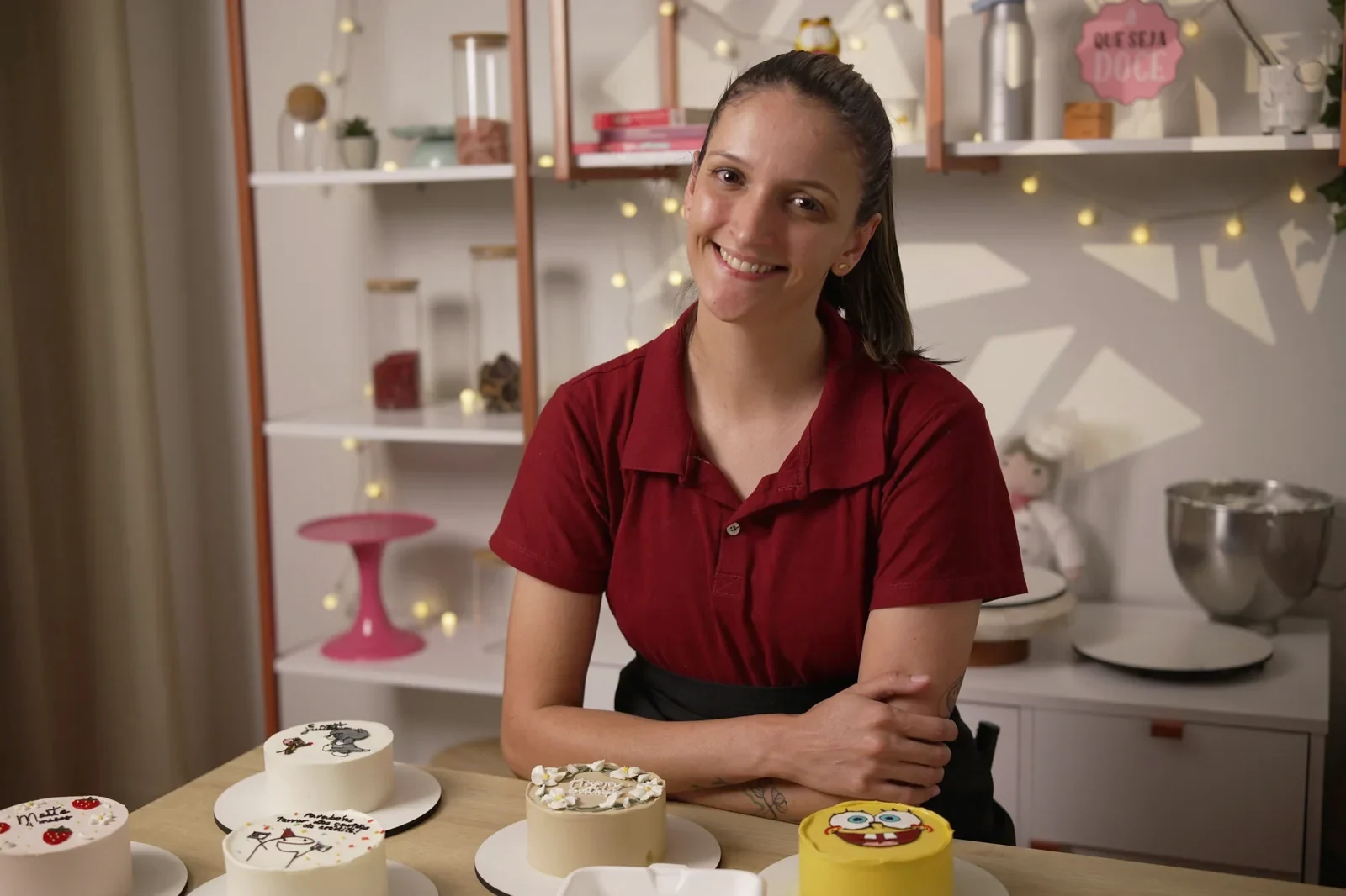 Amanda sorrindo na confeitaria, diante de nove modelos de bentô cakes